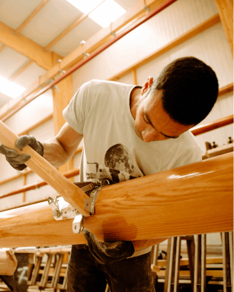 Mâts de voiliers en bois fabriqués artisanalement par un artisan penché au dessus dus mât en bois et tenant un espars en bois dans la main droite.