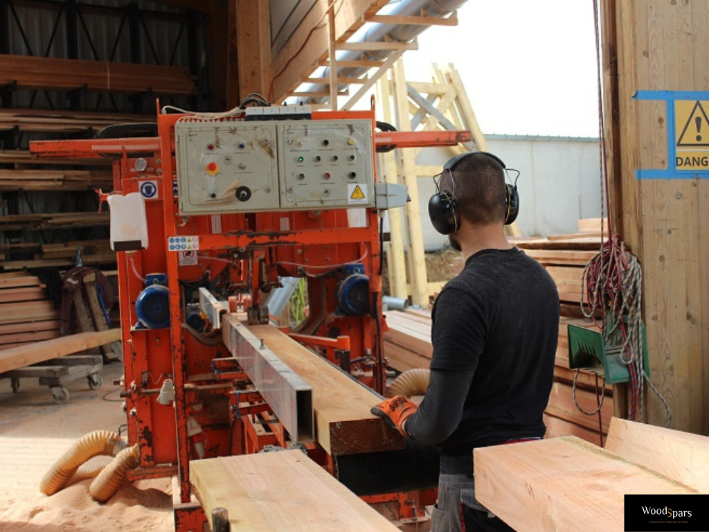 Artisan menuisier de Woodspars, équipé de protections auditives, usinant une poutre en bois massif sur une machine professionnelle dans l'atelier de l'entreprise.