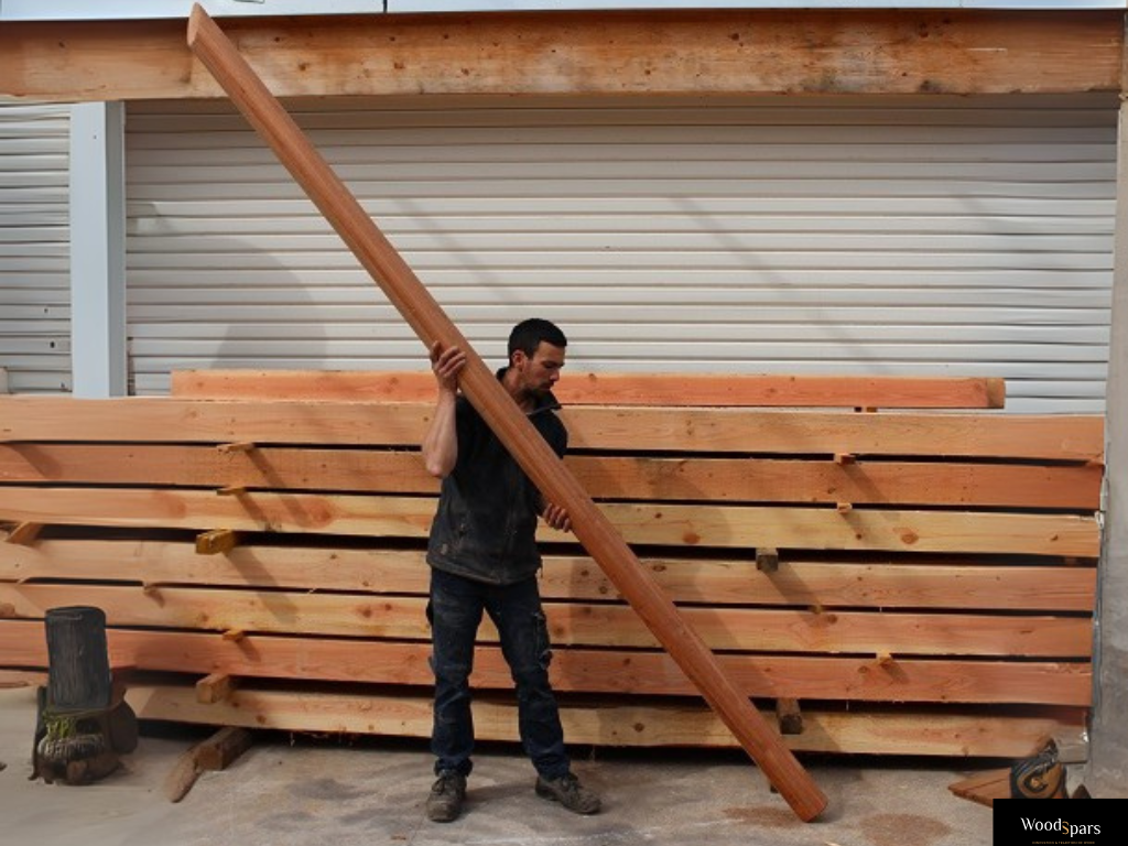 Artisan Woodspars transportant sur son épaule un mât rond pour voile d'ombrage en bois massif dans l'atelier, devant une pile de poutres en bois.