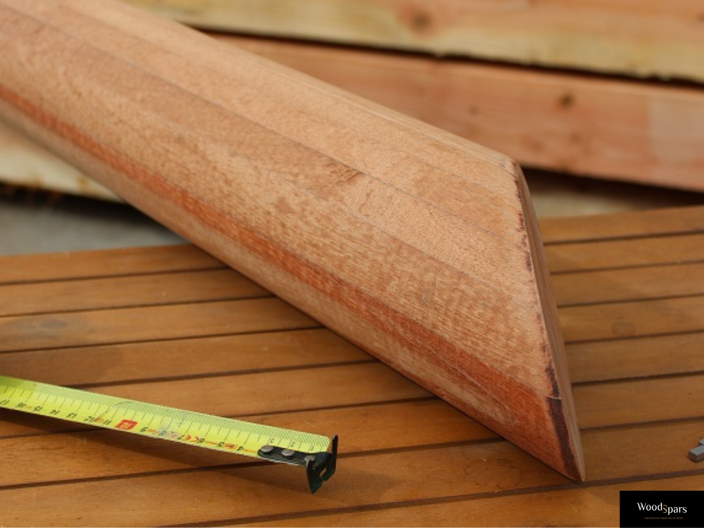 Gros plan sur la finition en pointe d'un mât pour voile d'ombrage en bois massif Woodspars, posé sur un établi à côté d'un mètre ruban symbolisant la fabrication sur mesure.