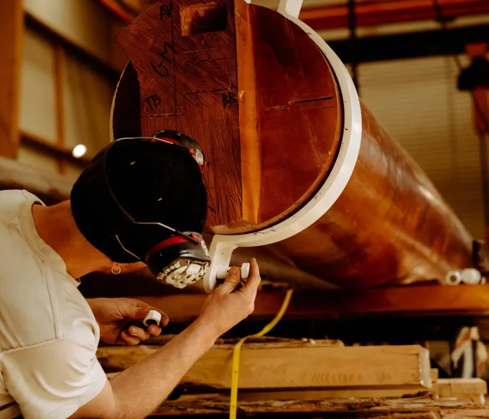 Artisan WoodSpars meulant la base d'un mât en bois lamellé collé posé sur tréteaux dans l'atelier de fabrication